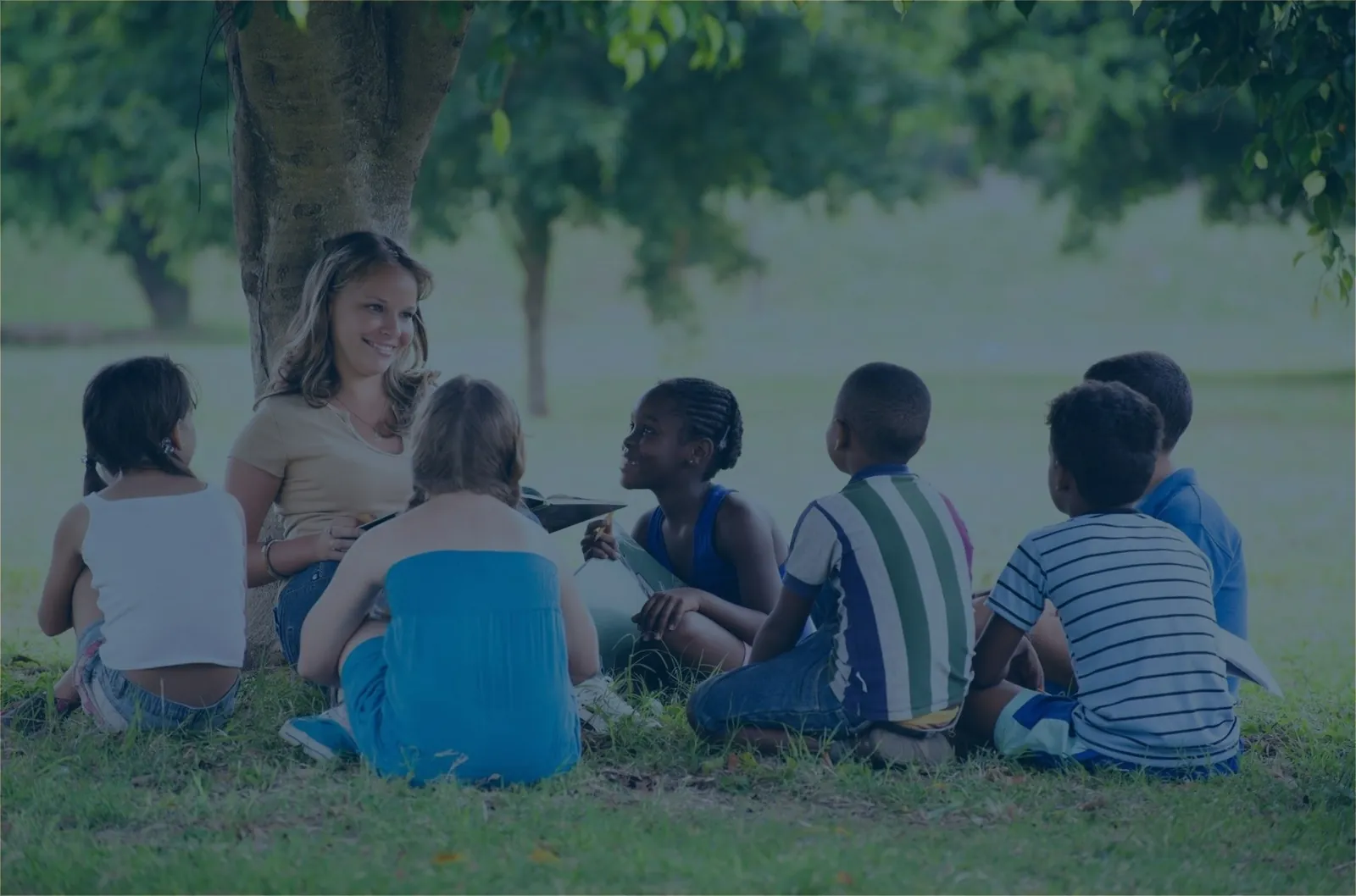 Teacher leading an outdoor circle lesson with children under a shady tree
